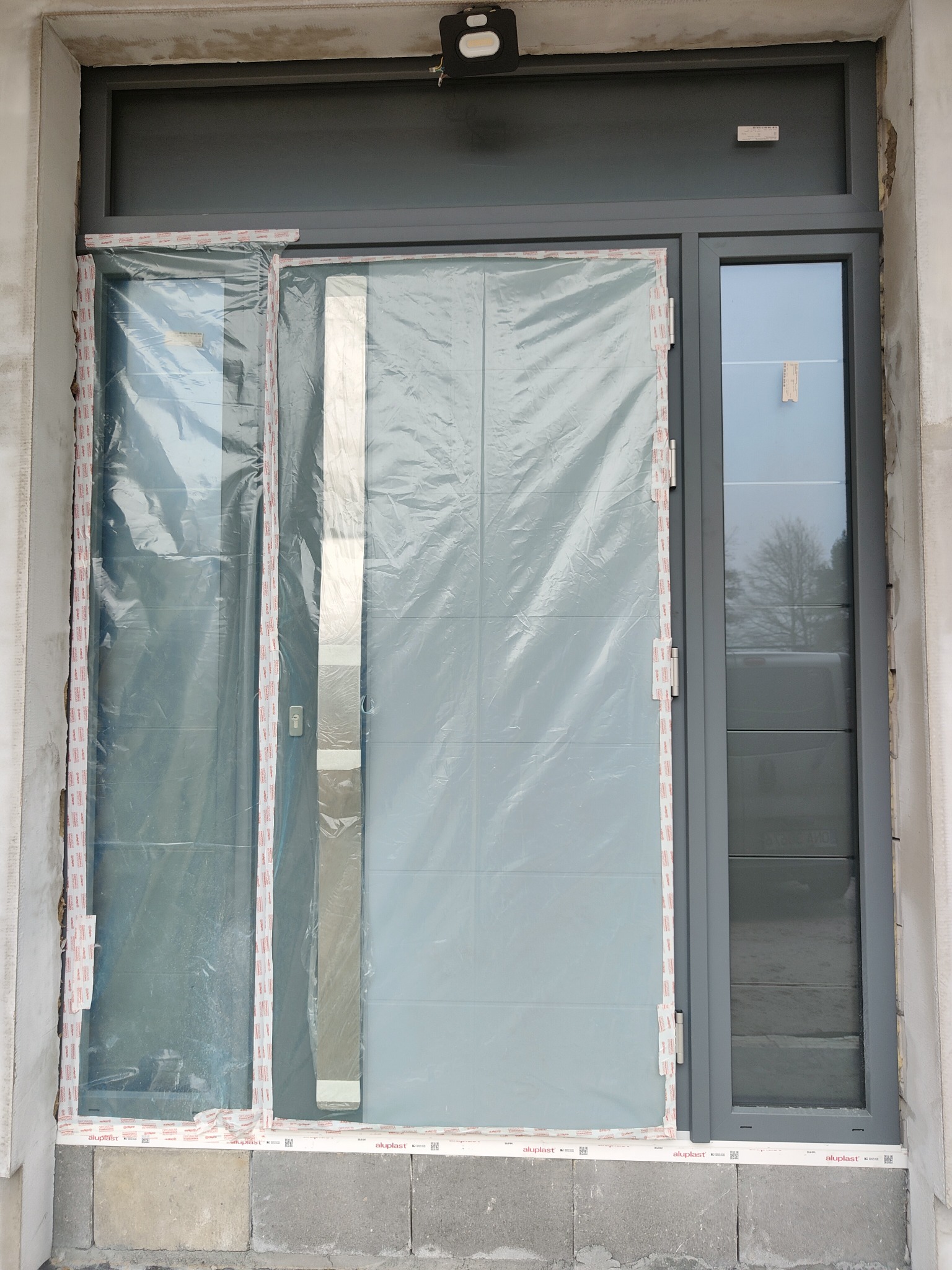 Szare drzwi wejściowe z bocznym oknem, zabezpieczone folią ochronną podczas montażu, widoczne ślady prac budowlanych na ścianie.