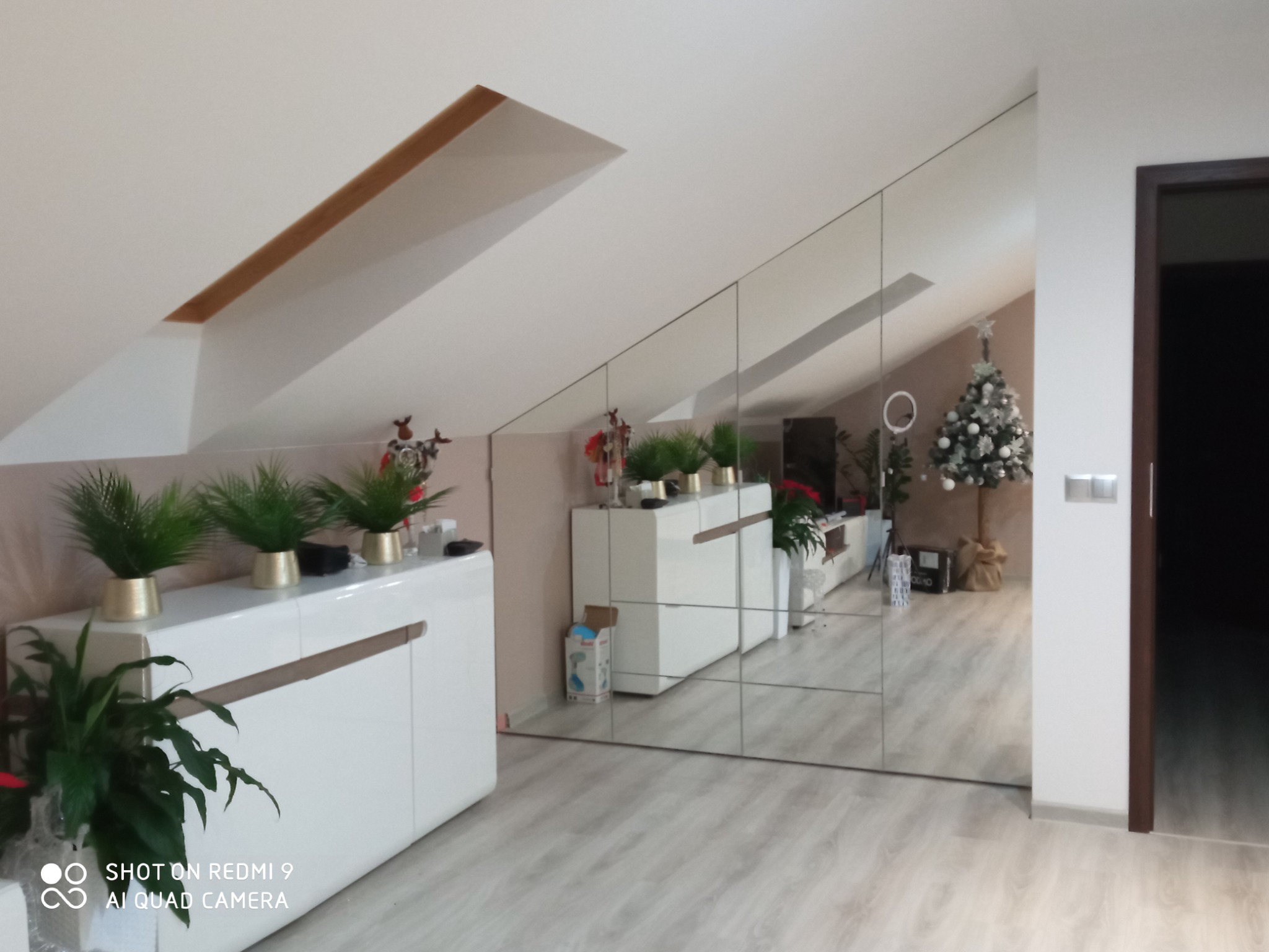 Skośny pokój na poddaszu z lustrzaną ścianą, odbijającą białe meble, choinkę i jasną podłogę.