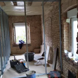 Montaż metalowej konstrukcji pod płytę karton gips na tle surowej ceglanej ściany w remontowanym pomieszczeniu z oknami.