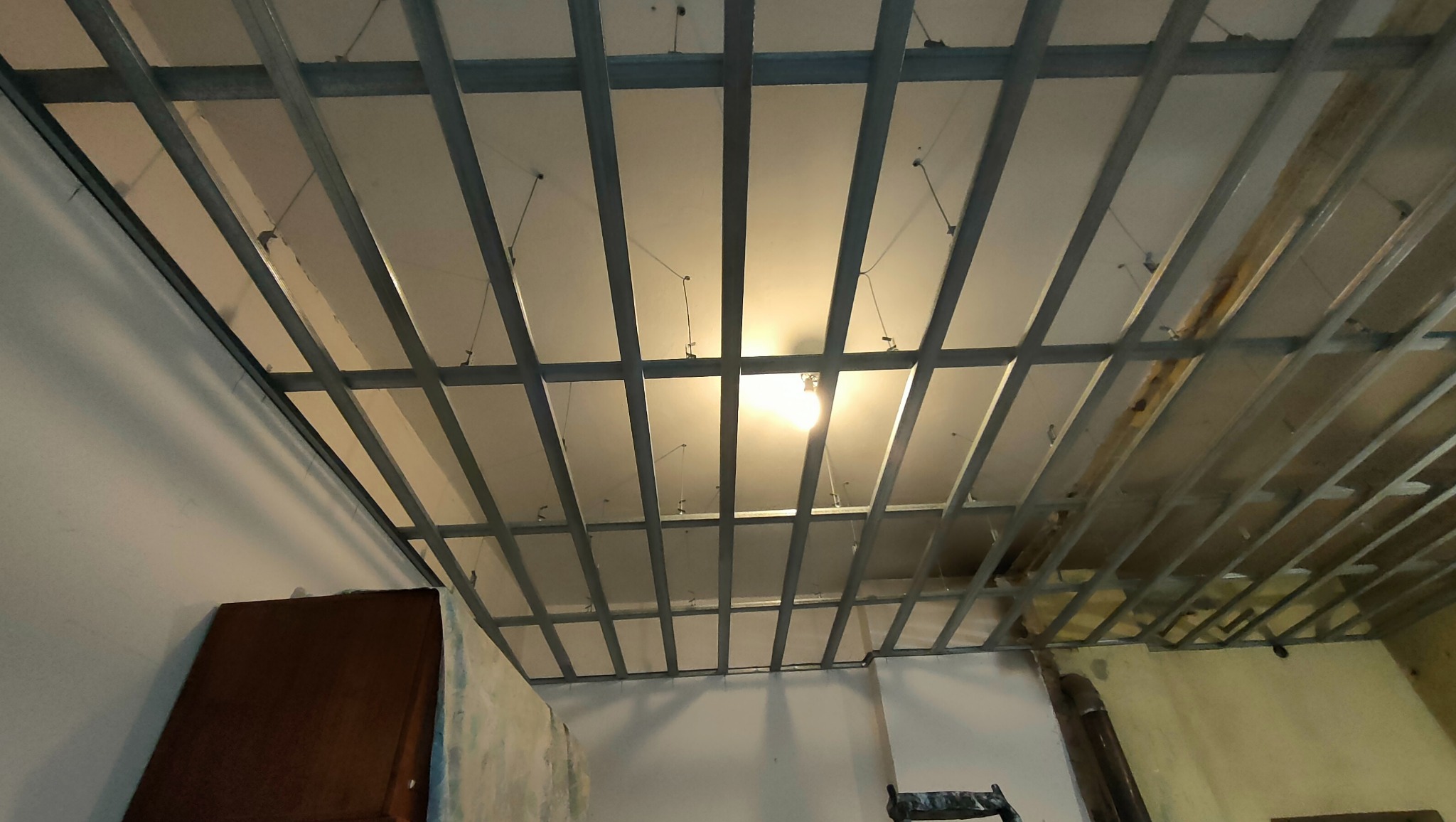 Metalowa konstrukcja sufitu podwieszanego z zamontowaną płytą gipsowo-kartonową, widok z dołu, widoczna instalacja elektryczna i oświetlenie tymczasowe.