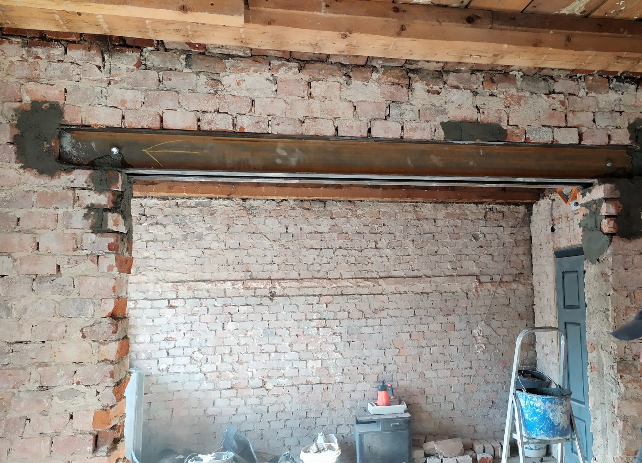 Wstawiony stalowy profil w ścianie z cegły, wzmocniony zaprawą, strzałka wskazująca kierunek, widoczne narzędzia i drabina.