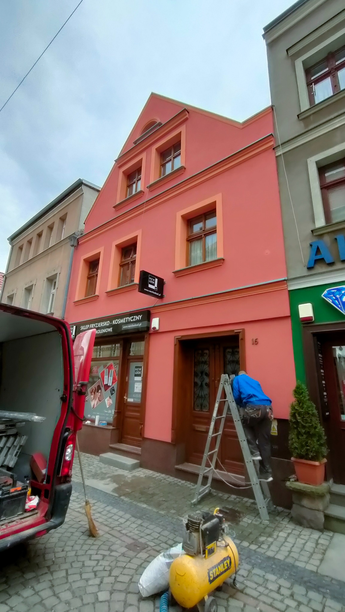 Pracownik na drabinie przy wejściu do budynku, prawdopodobnie wykonujący prace wykończeniowe elewacji, z widocznym kompresorem Stanley i otwartym busem z narzędziami w tle.