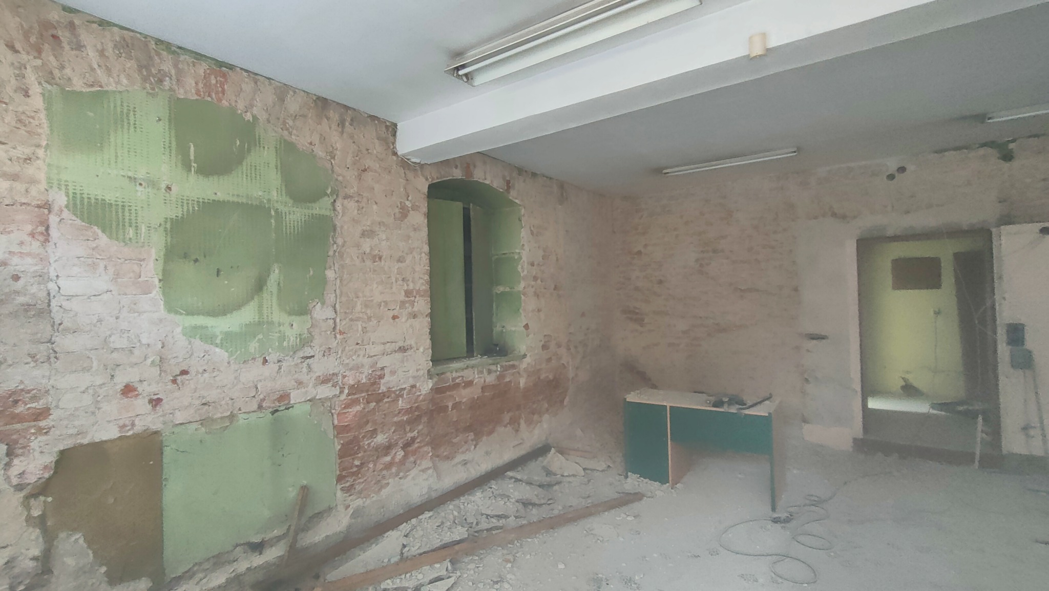 Wnętrze w trakcie remontu: odsłonięta cegła, ślady po zielonym podkładzie, fragmenty gruzu na podłodze, surowe okno i biurko w rogu, widoczne instalacje elektryczne.