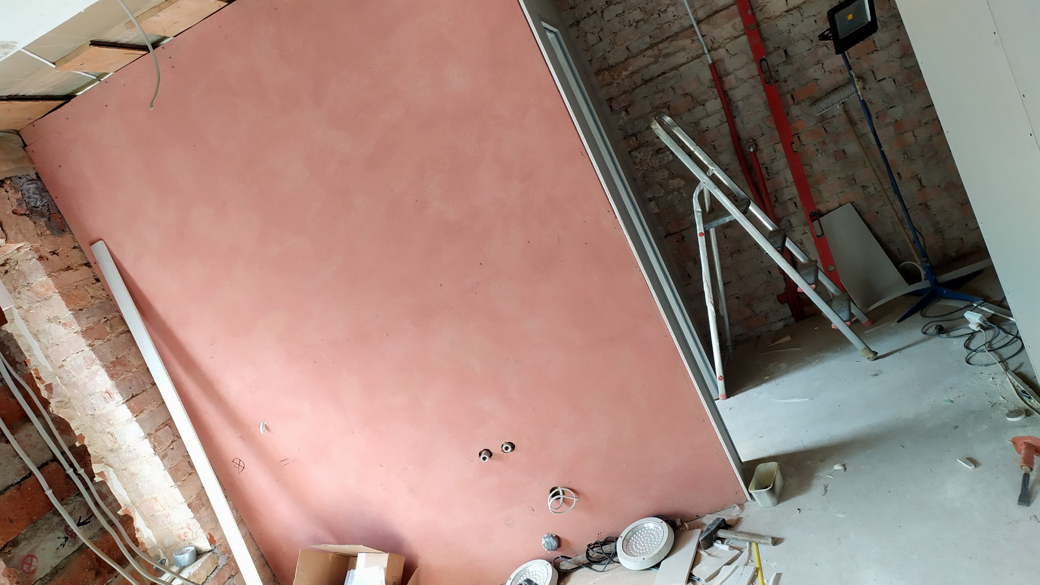 Różowa, świeżo wyszpachlowana ściana w trakcie remontu, widoczne puszki elektryczne, fragment ceglanej ściany i drabina.