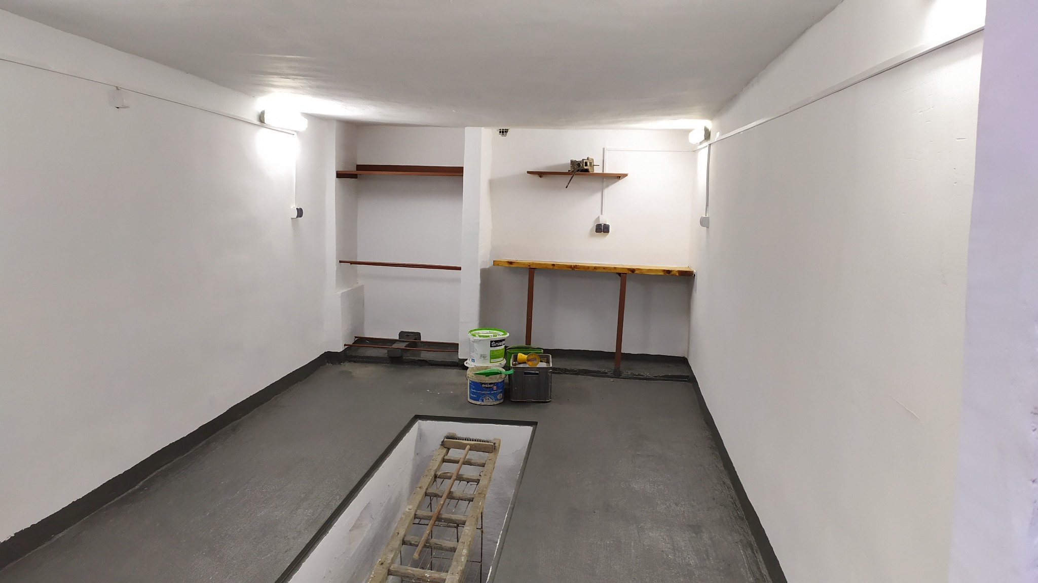 Wnętrze piwnicy po malowaniu, szare podłogi i białe ściany, widoczne puszki z farbą, drabina w kanale podłogowym i prowizoryczne półki