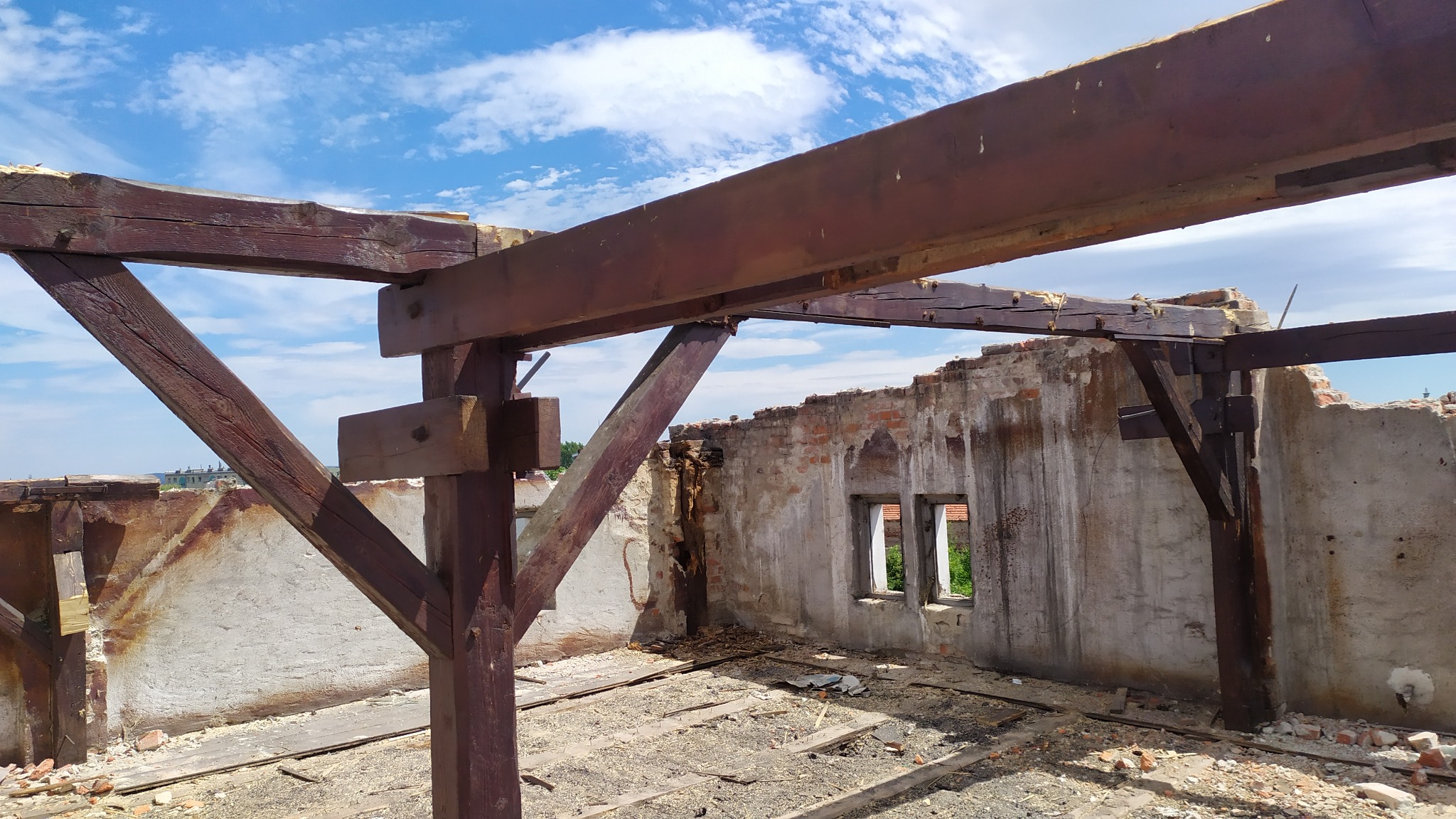 Wnętrze zniszczonego budynku z odsłoniętą drewnianą konstrukcją dachu i fragmentami ścian z widocznymi oknami, ukazujące stan przed remontem lub w trakcie rozbiórki.