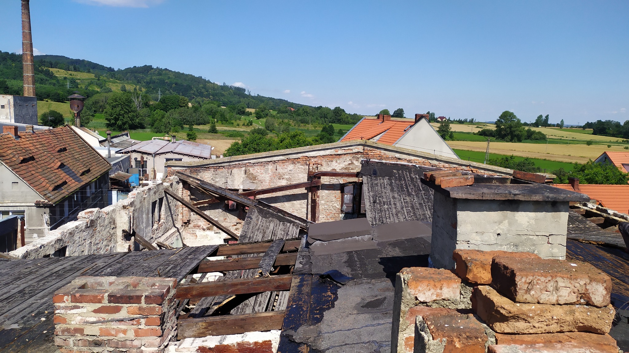 Widok z góry na częściowo zrujnowany budynek z cegły i drewna, z widocznymi elementami konstrukcyjnymi, kominami i okoliczną zabudową oraz polami w tle, w słoneczny dzień.