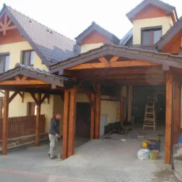 Malowanie drewnianej konstrukcji wiaty przydomowej, widoczne narzędzia i drabina. Dach domu z brązową dachówką w tle.