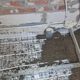 Montaż ogrzewania podłogowego: widok na rozłożoną siatkę z rurami, częściowo pokrytą mokrą wylewką, z widoczną ścianą z cegły i instalacją sanitarną przy suficie.