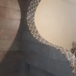 Ściana wyłożona płytkami ceramicznymi w odcieniach szarości i beżu, z dekoracyjnym, półokrągłym lustrem z kryształkami, tworzącymi falisty kształt.