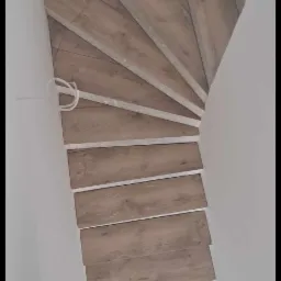 Montaż drewnopodobnych płytek na schodach spiralnych, widok z góry, widoczne narzędzie glazurnicze i kabel elektryczny wystający ze ściany.