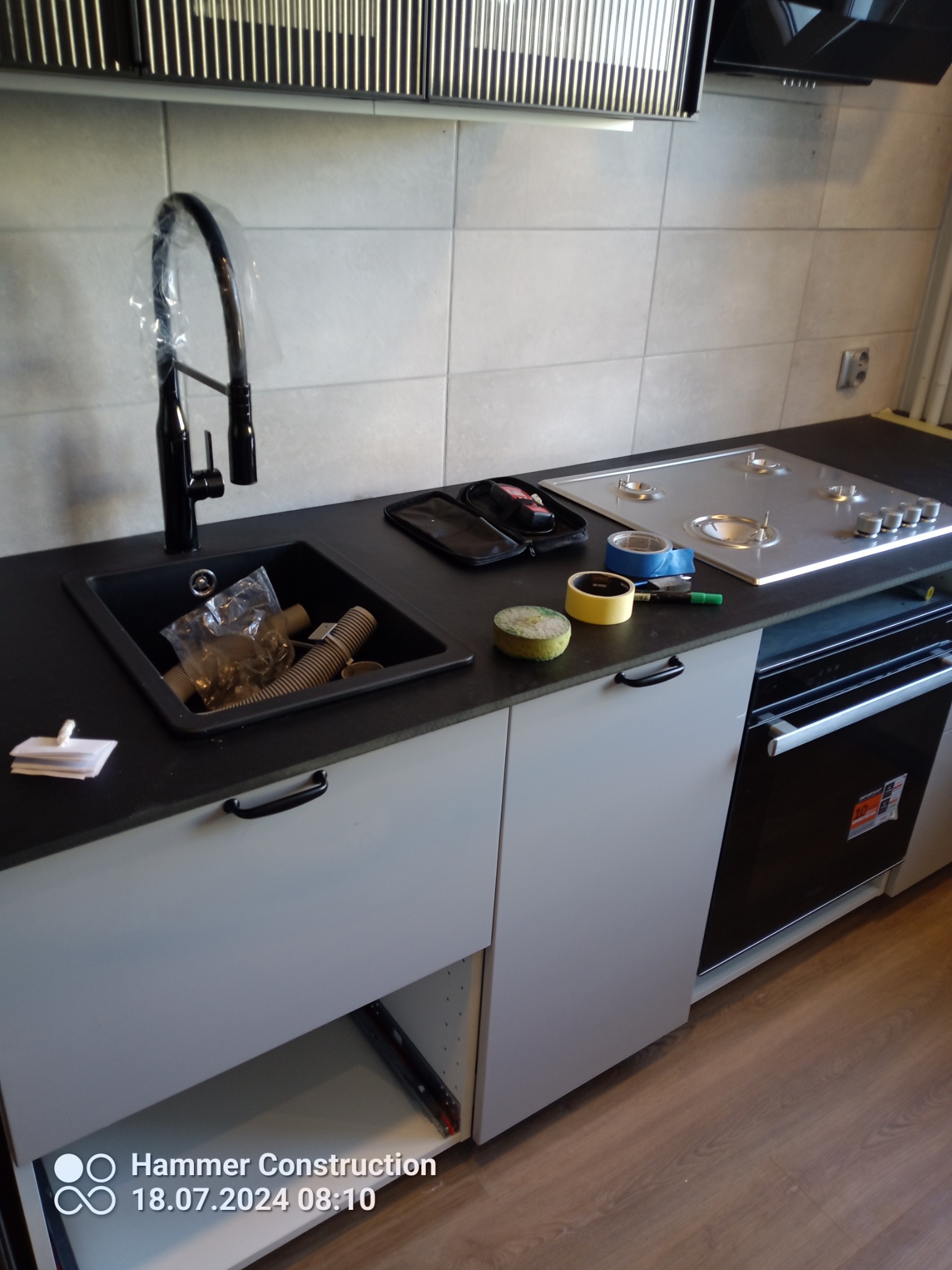 Montaż czarnej baterii kuchennej z folią ochronną nad czarnym zlewozmywakiem, obok płyta gazowa z taśmą malarską, widoczne narzędzia i akcesoria, otwarte szafki kuchenne.