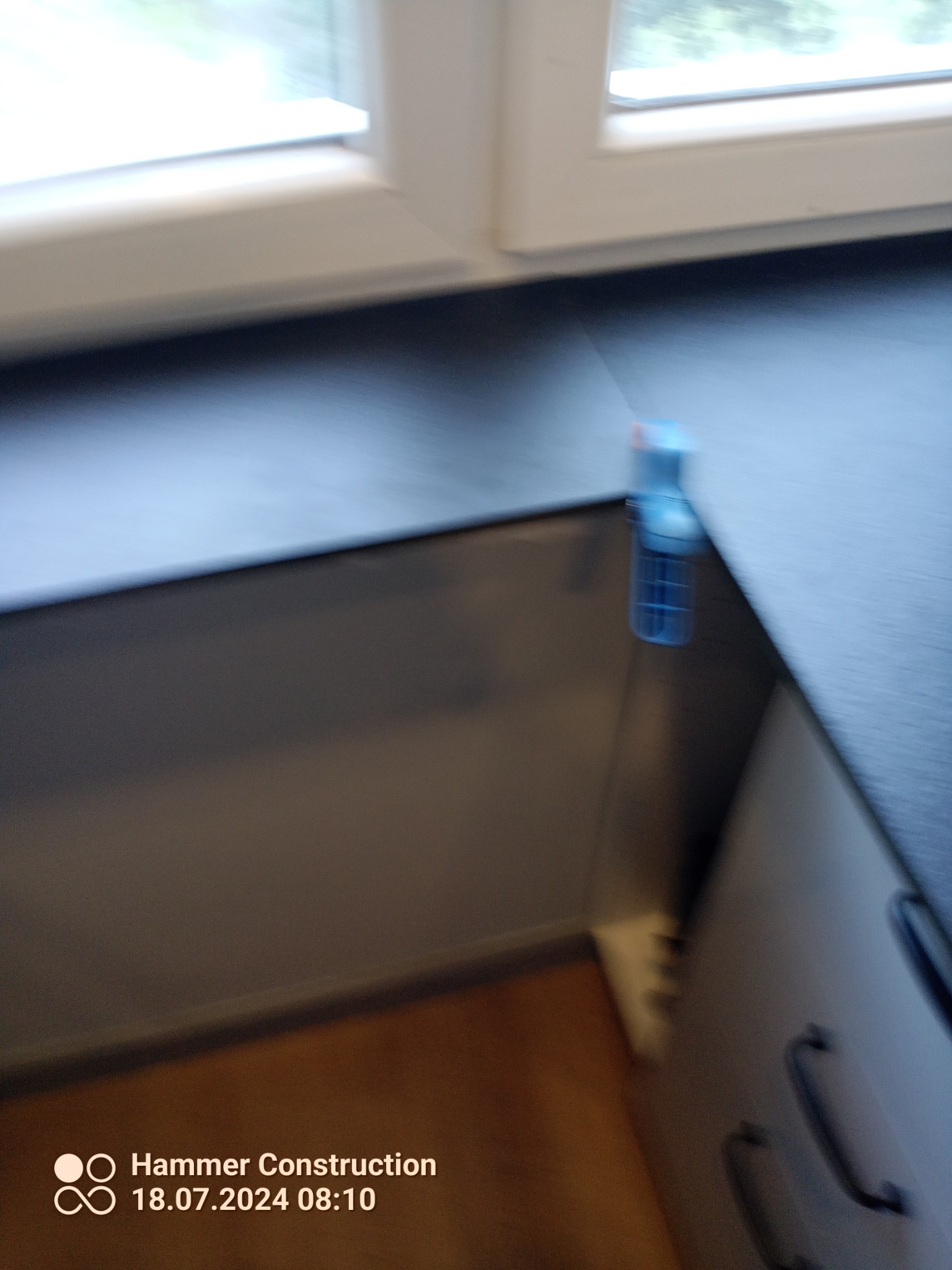 Rozmazane ujęcie fragmentu blatu kuchennego przy oknie, z widoczną butelką środka czyszczącego i fragmentem szafki z uchwytami.