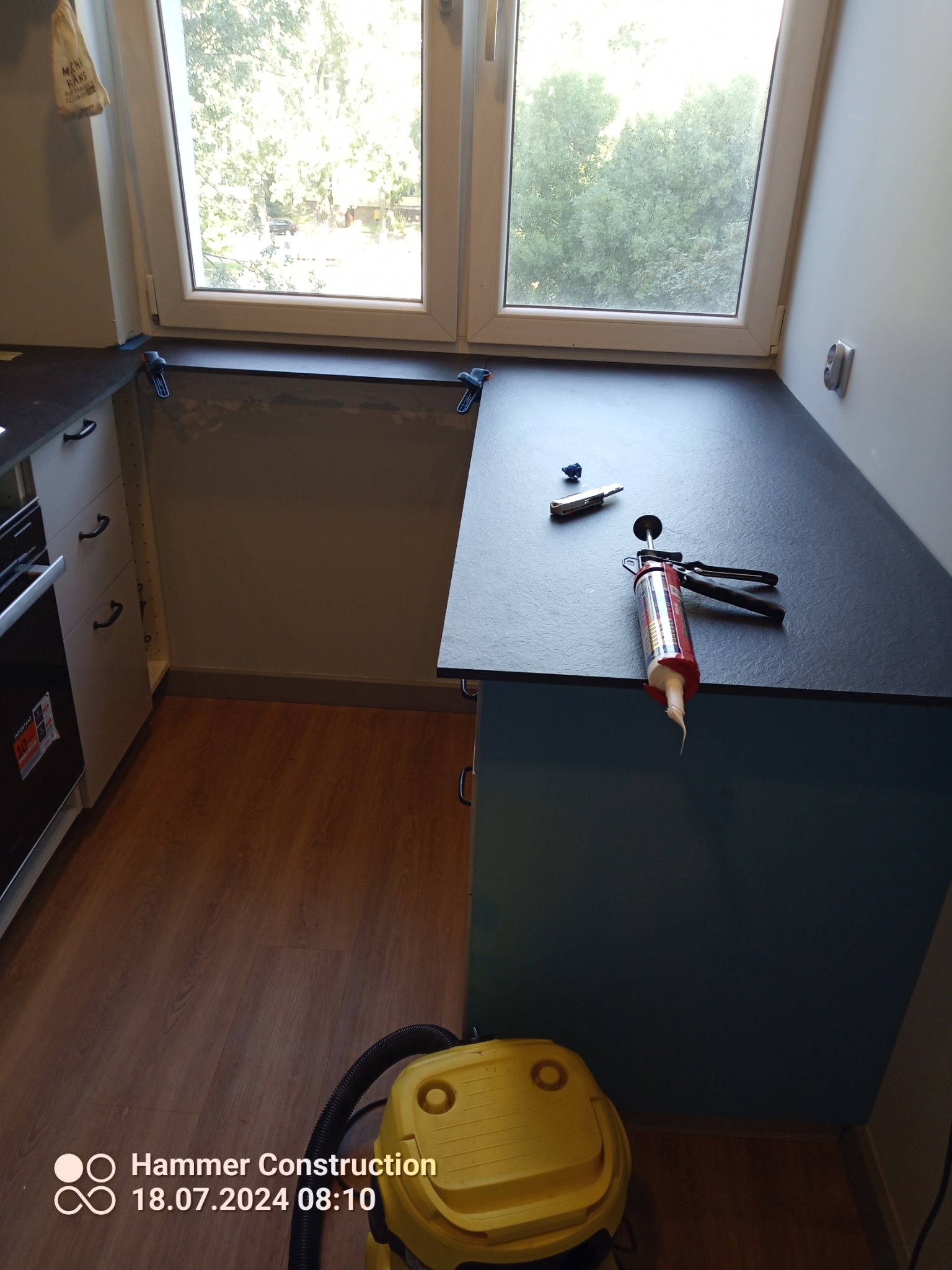 Montaż blatu kuchennego: widoczna tuba z silikonem, nóż do tapet, niebieskie zaciski stolarskie przy oknie oraz odkurzacz na podłodze w trakcie prac wykończeniowych.