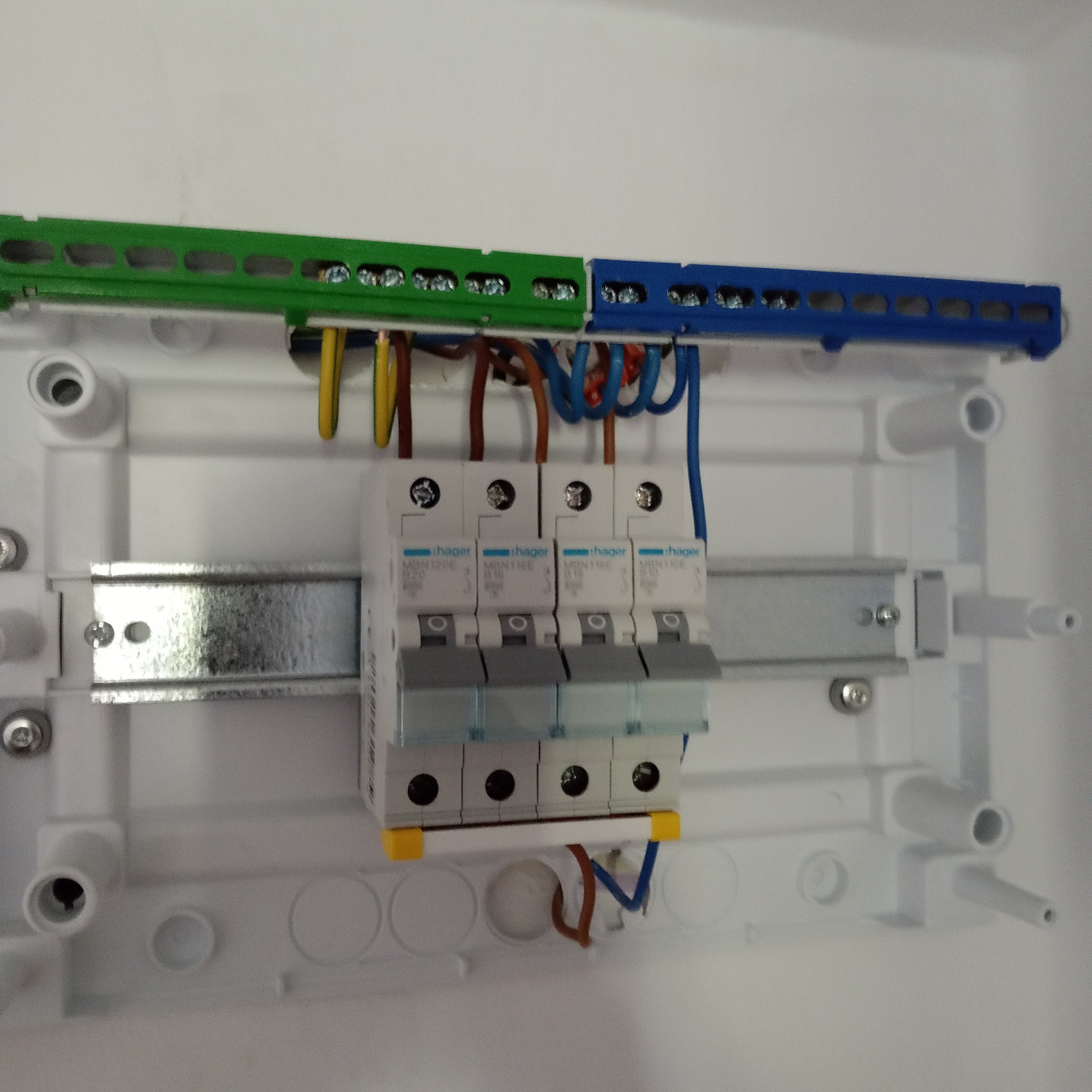 Rozdzielnica elektryczna z zamontowanymi bezpiecznikami Hager, widoczne przewody w kolorach żółtym, brązowym i niebieskim oraz szyny DIN w kolorach zielonym i niebieskim.