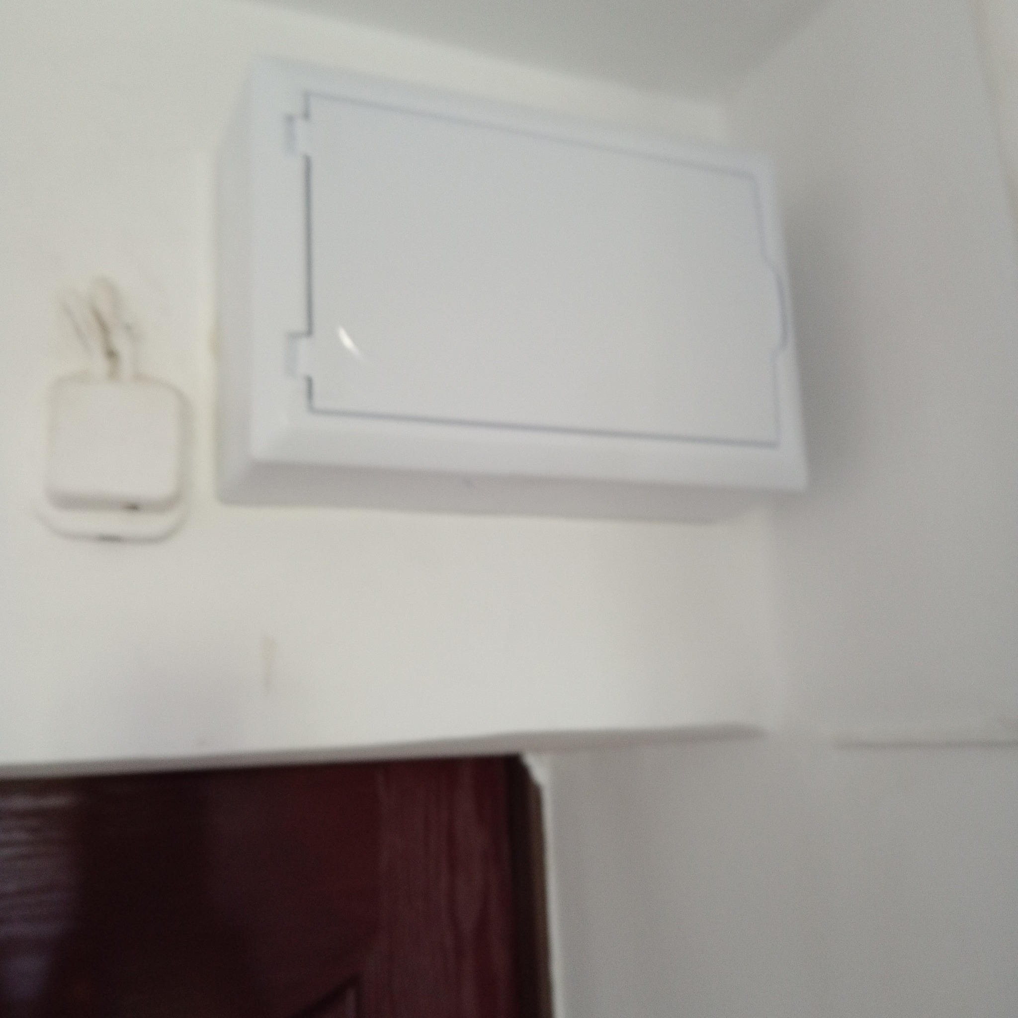 Biała skrzynka elektryczna z widocznym osprzętem na ścianie, obok fragmentu futryny drzwiowej.