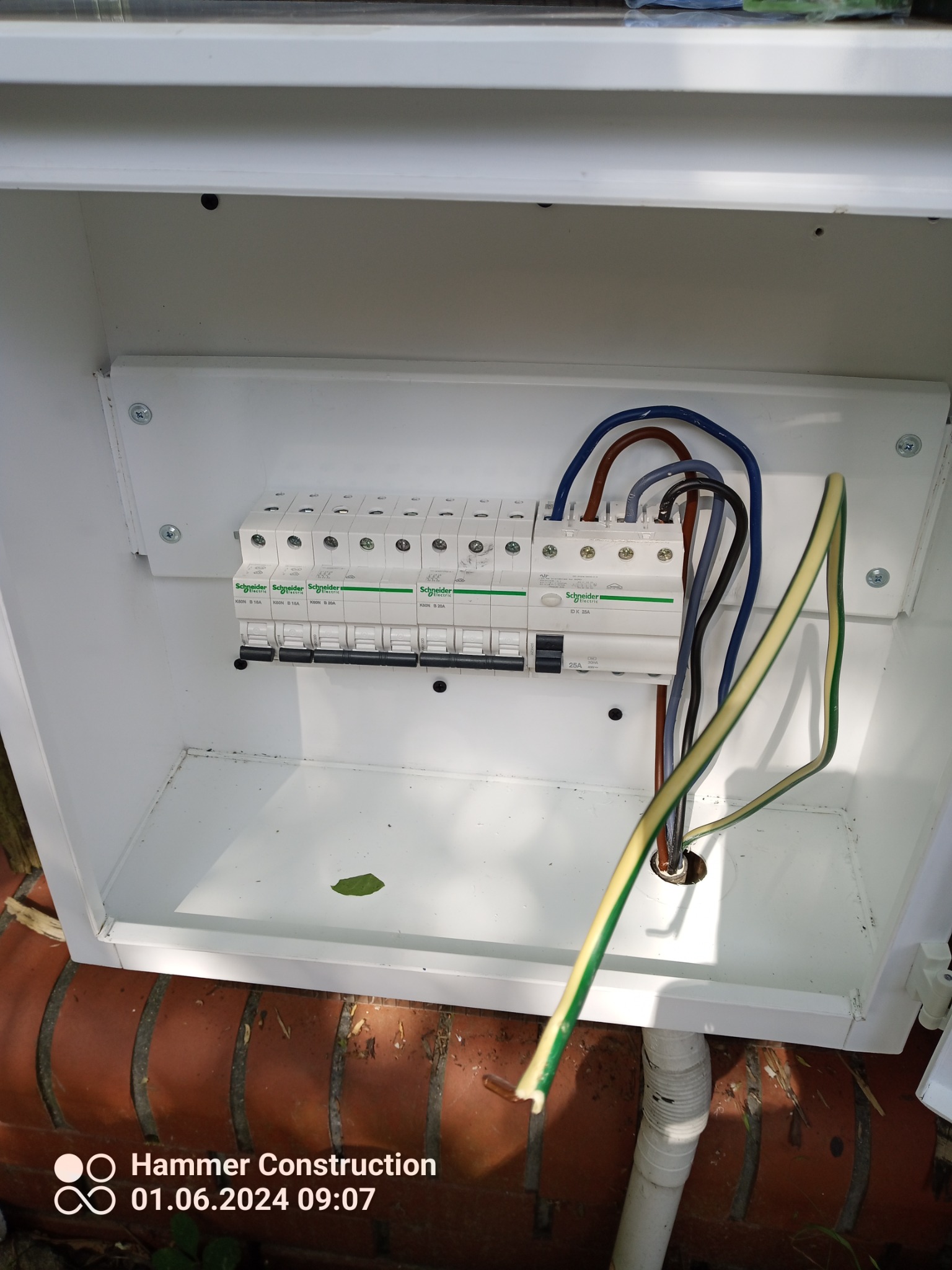 Biała skrzynka elektryczna z zamontowanymi bezpiecznikami Schneider, widoczne przewody w różnych kolorach. Skrzynka umieszczona na ceglanej podstawie.