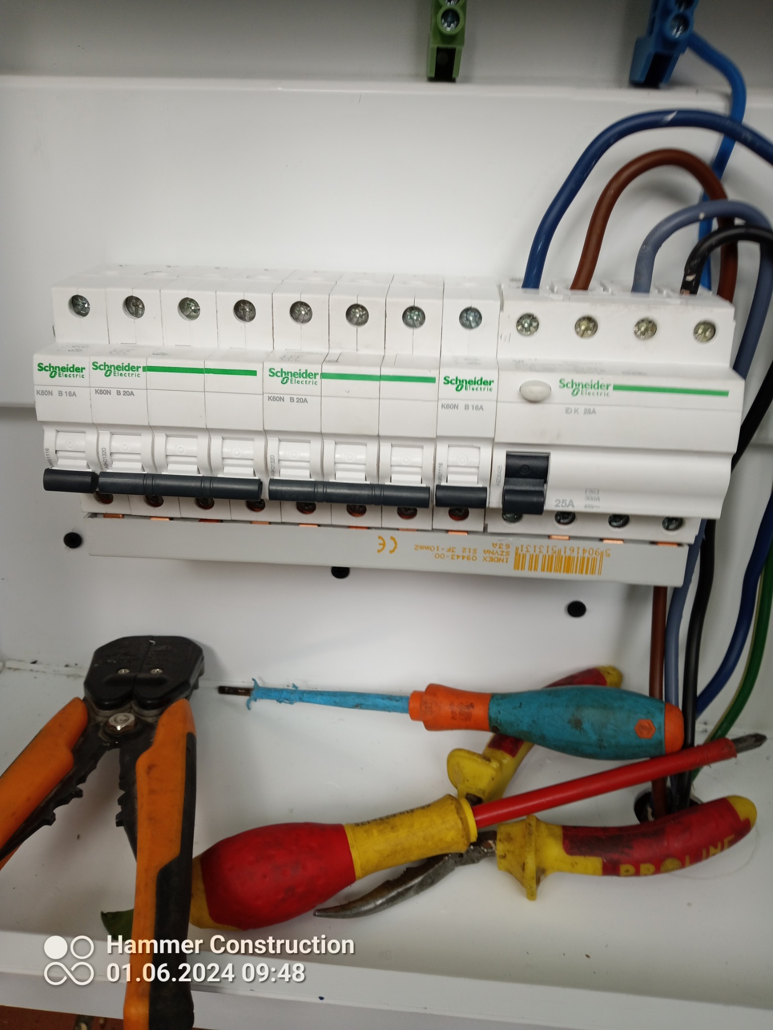 Rozdzielnica elektryczna Schneider Electric z zamontowanymi bezpiecznikami, widoczne przewody i narzędzia: kombinerki, śrubokręty z niebieską i czerwoną rękojeścią.