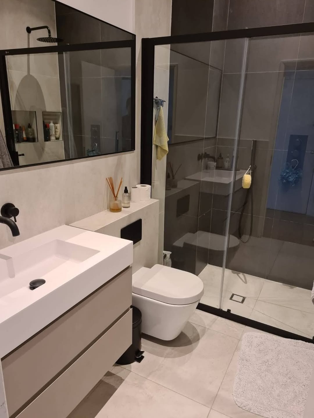 Nowoczesna łazienka z kabiną prysznicową ze szklanymi drzwiami przesuwnymi w czarnej ramie, umywalką nablatową na szafce w odcieniach beżu, wiszącą toaletą i dużym lustrem.