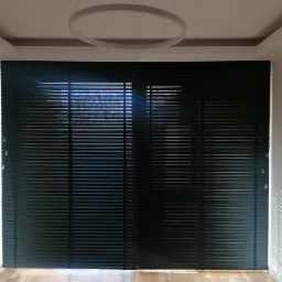 Czarne żaluzje poziome drewniane z widocznymi listwami, zamontowane w oknie, oświetlone światłem słonecznym wpadającym przez szpary, z parkietem na podłodze i nowoczesną okrągłą lampą sufitową.