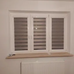 Trzy okna z zamontowanymi roletami poziomymi dzień i noc w kolorze szaro-białym, widoczne na tle białej ściany i grzejnika.