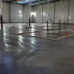 Wnętrze hali z wylaną, mokrą posadzką betonową, z wystającymi prętami zbrojeniowymi i elementami szalunku. Widoczne odbicia światła na gładkiej powierzchni betonu.