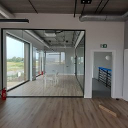 Sawa - Nowoczesne biuro z przeszkloną ścianą działową, widok na zewnątrz, drewniana podłoga, surowy betonowy sufit z instalacjami. Gaśnica przy ścianie.