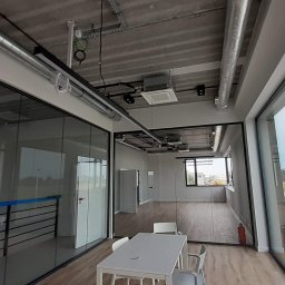 Sawa - Wnętrze biurowe z przeszklonymi ścianami działowymi, widoczne instalacje pod sufitem, biurko z krzesłami. Surowy, nowoczesny design.
