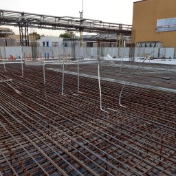 Sawa - Zbrojenie fundamentów pod budynek przemysłowy, widok z góry na stalową siatkę. W tle pracownicy w odblaskowych ubraniach i elementy infrastruktury przemysłowej.