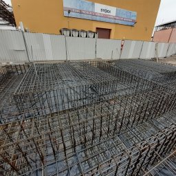 Sawa - Zbrojenie fundamentów pod budynek z widoczną konstrukcją stalową, czarną folią izolacyjną i częścią budynku w tle. Kąt ujęcia z góry, oświetlenie dzienne.