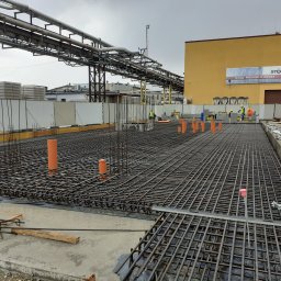 Sawa - Zbrojenie fundamentów z rurami instalacyjnymi na budowie, w tle widoczny budynek z reklamą. Stalowe rury nad placem budowy.