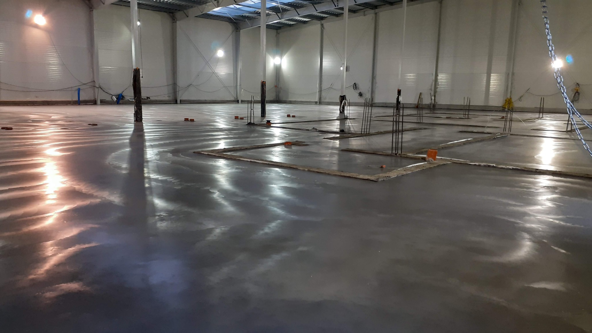 Wnętrze hali z wylaną, mokrą posadzką betonową, z wystającymi prętami zbrojeniowymi i elementami szalunku. Widoczne odbicia światła na gładkiej powierzchni betonu.