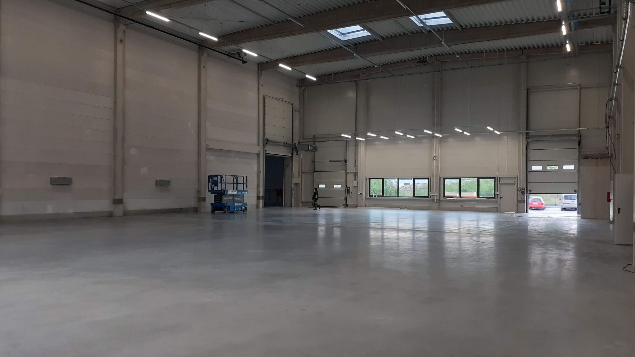 Wnętrze hali produkcyjnej z betonową posadzką, widoczne bramy segmentowe i podnośnik koszowy. Światło dzienne wpada przez okna i świetliki dachowe.