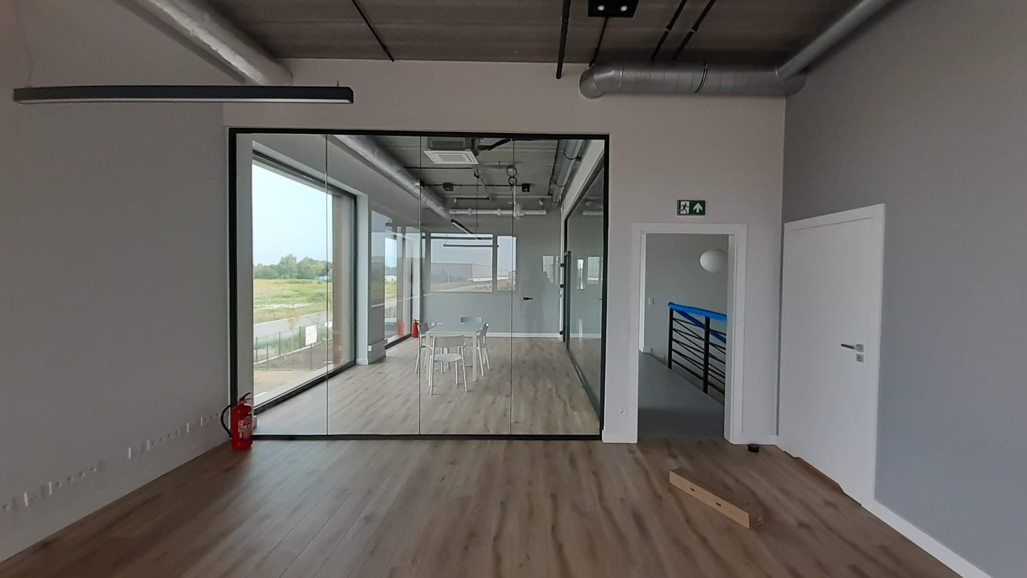 Nowoczesne biuro z przeszkloną ścianą działową, widok na zewnątrz, drewniana podłoga, surowy betonowy sufit z instalacjami. Gaśnica przy ścianie.