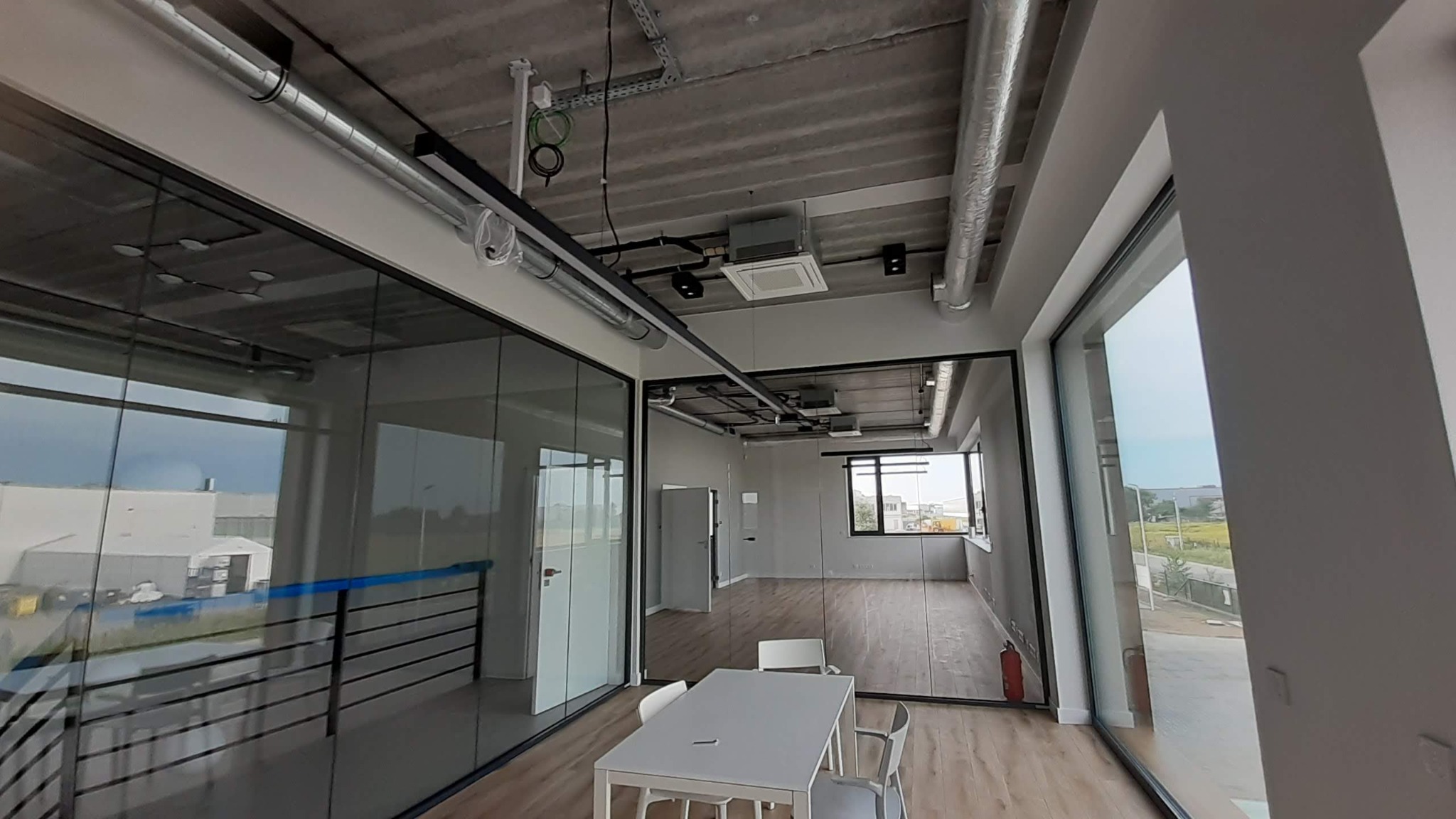 Wnętrze biurowe z przeszklonymi ścianami działowymi, widoczne instalacje pod sufitem, biurko z krzesłami. Surowy, nowoczesny design.