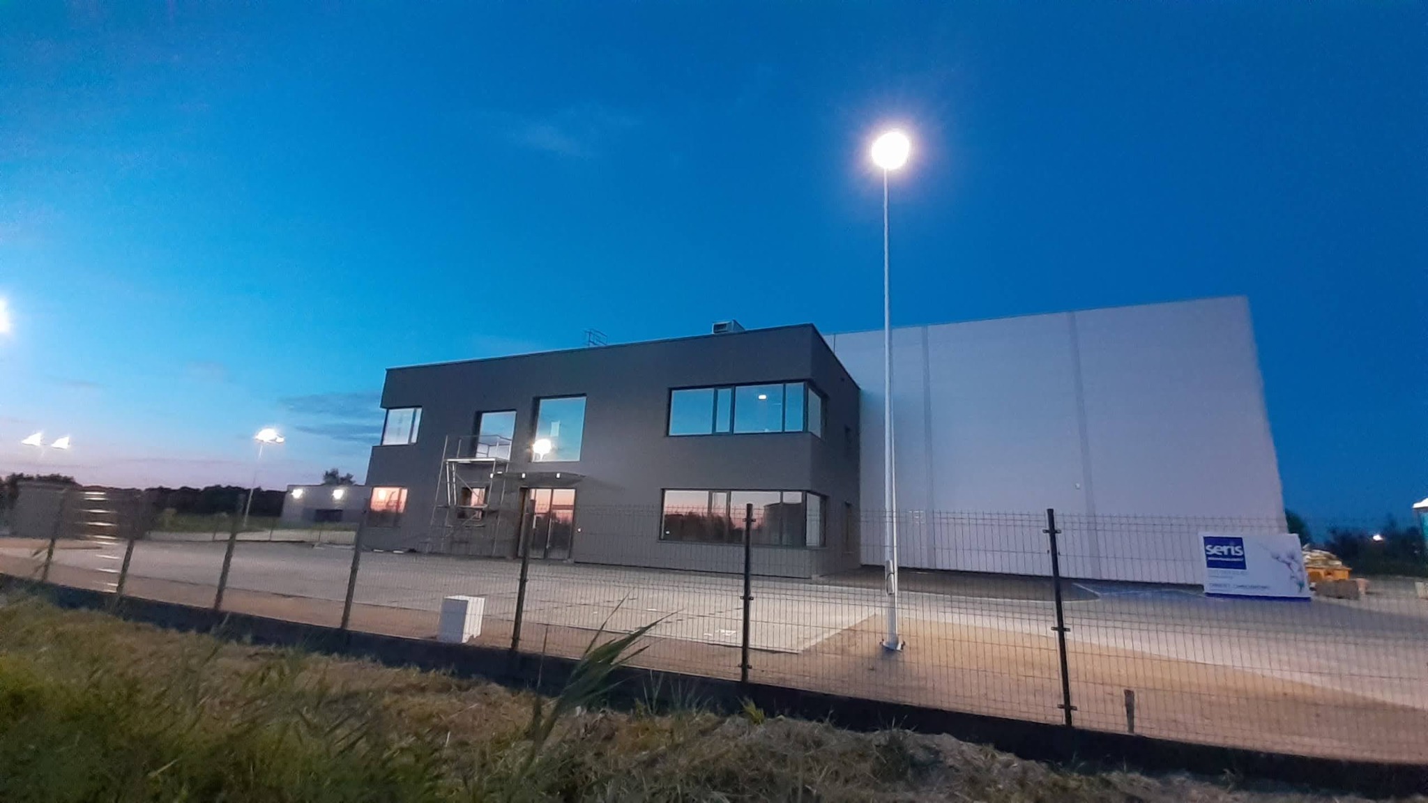 Nowoczesny budynek biurowy z szarą elewacją i dużymi oknami, oświetlony lampami ulicznymi, widziany o zmierzchu za ogrodzeniem.