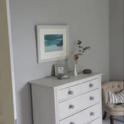 Jasnoszary pokój po odświeżeniu: ściany pomalowane na jednolity kolor, biała komoda z dekoracjami, obrazek na ścianie i fotel w tle.