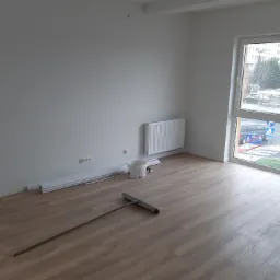 Pusty pokój po remoncie z jasnymi panelami podłogowymi, świeżo pomalowanymi ścianami na biało, grzejnikiem, listwami przypodłogowymi, wiadrem po farbie i narzędziem do rozprowadzania wylewki...