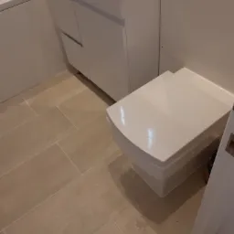 Nowoczesna, podwieszana toaleta w łazience z beżowymi płytkami podłogowymi, obok umywalka z szafką i fragment wanny.