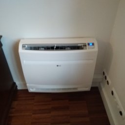 JKB KLIMA KRZYSZTOF BUSZYŃSKI - Biały klimatyzator podłogowy LG z niebieską diodą włączony, zamontowany w rogu pokoju obok ciemnej komody i listwy przypodłogowej z gniazdkami, widoczny parkiet.