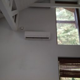 JKB KLIMA KRZYSZTOF BUSZYŃSKI - Biały klimatyzator ścienny zamontowany w pokoju z oknami i drewnianą konstrukcją sufitu, widok na zewnątrz przez okno.