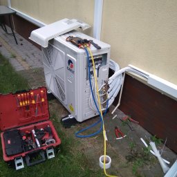 JKB KLIMA KRZYSZTOF BUSZYŃSKI - Jednostka zewnętrzna klimatyzatora marki Gree, z otwartą obudową, podłączona do przewodów, narzędzia pomiarowe na górze, obok otwarta skrzynka z narzędziami na trawie, widoczne elementy instalacji...