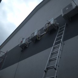 JKB KLIMA KRZYSZTOF BUSZYŃSKI - Zewnętrzne jednostki klimatyzacyjne zamontowane rzędem na dwukolorowej elewacji budynku, z aluminiową drabiną opartą o ścianę i fragmentem rusztowania w kadrze.