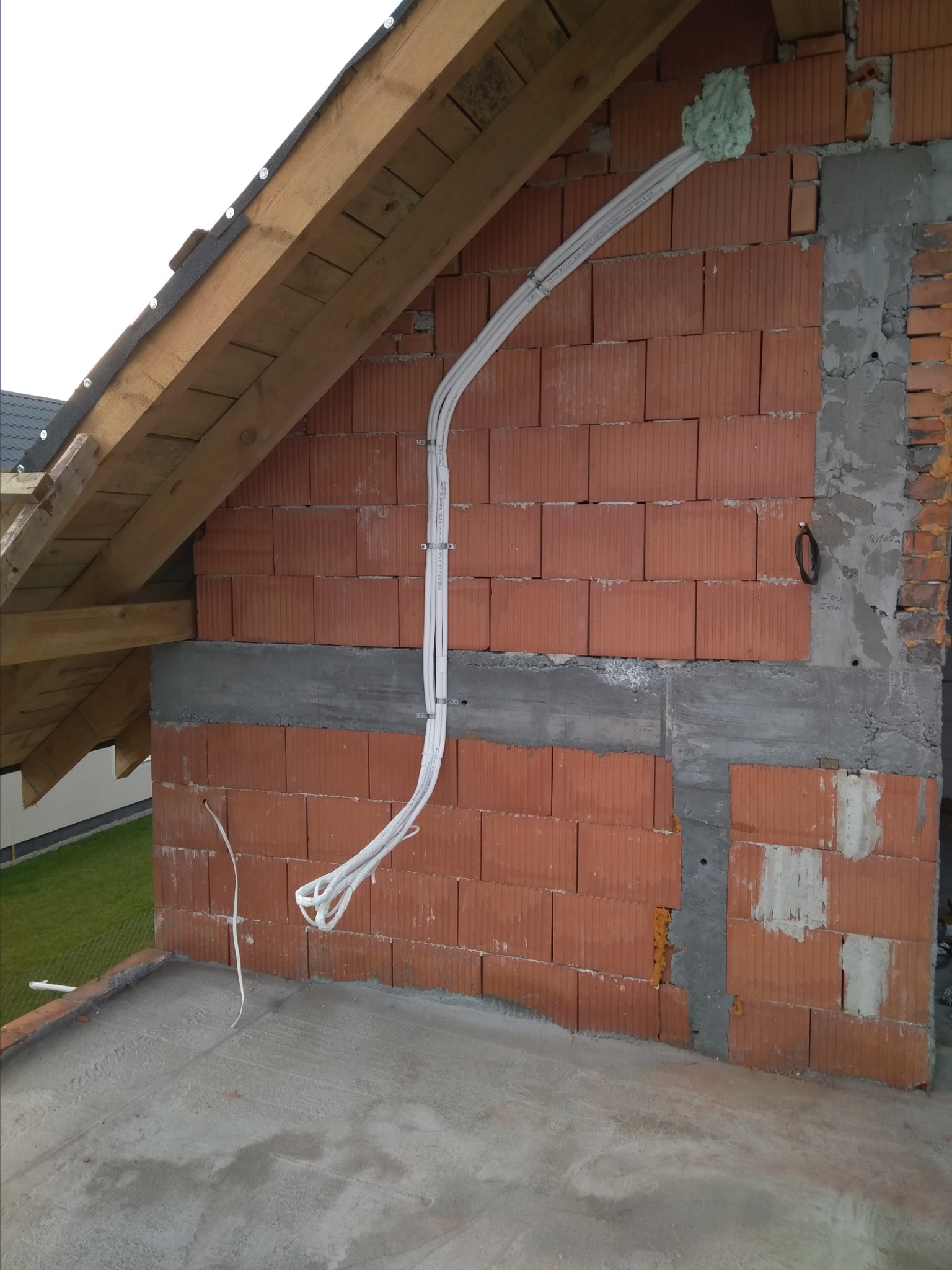 Montaż przewodów wentylacyjnych na ceglanej ścianie poddasza, zabezpieczone pianką montażową, widoczne drewniane elementy konstrukcji dachu.