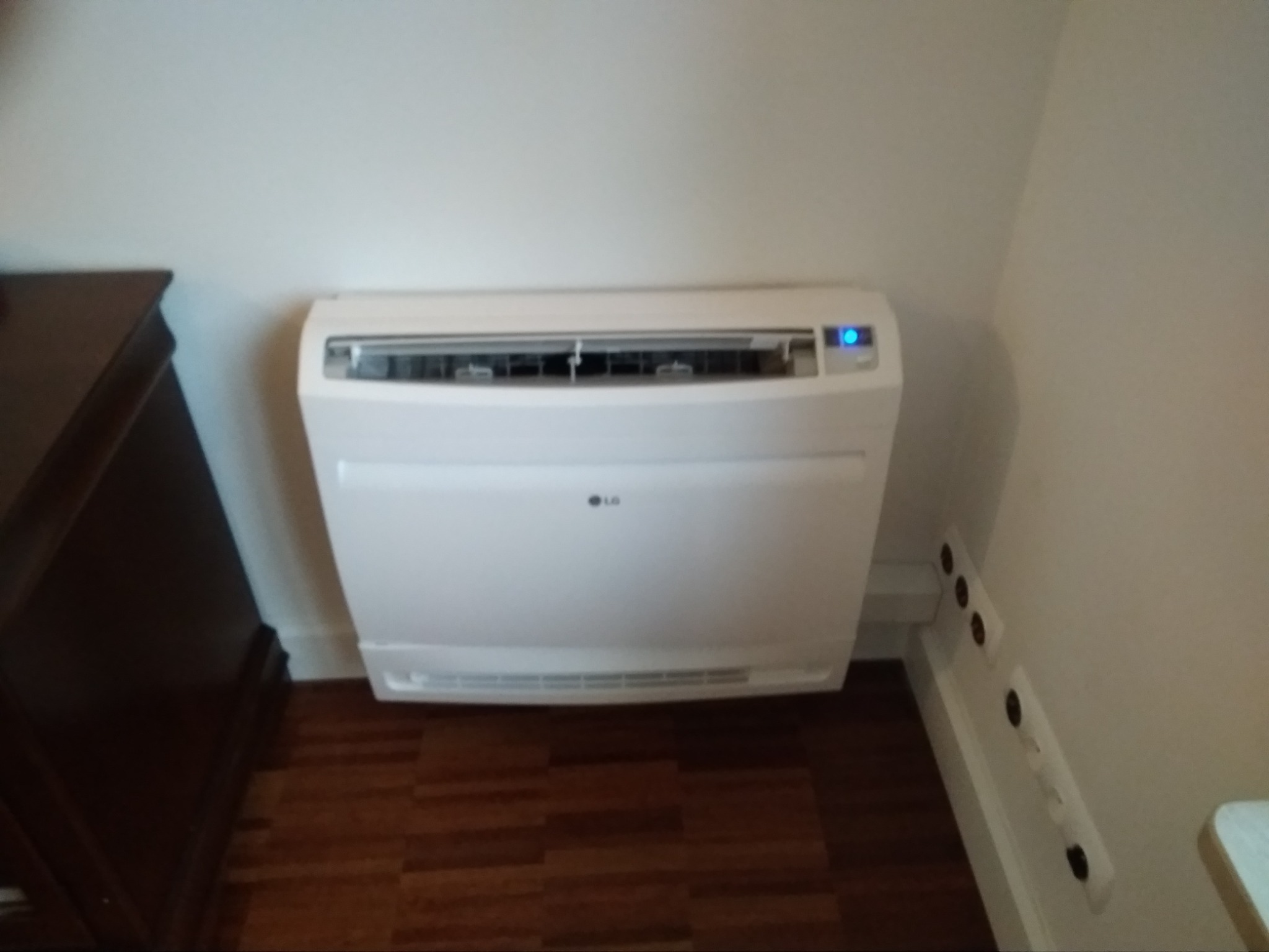 Biały klimatyzator podłogowy LG z niebieską diodą włączony, zamontowany w rogu pokoju obok ciemnej komody i listwy przypodłogowej z gniazdkami, widoczny parkiet.