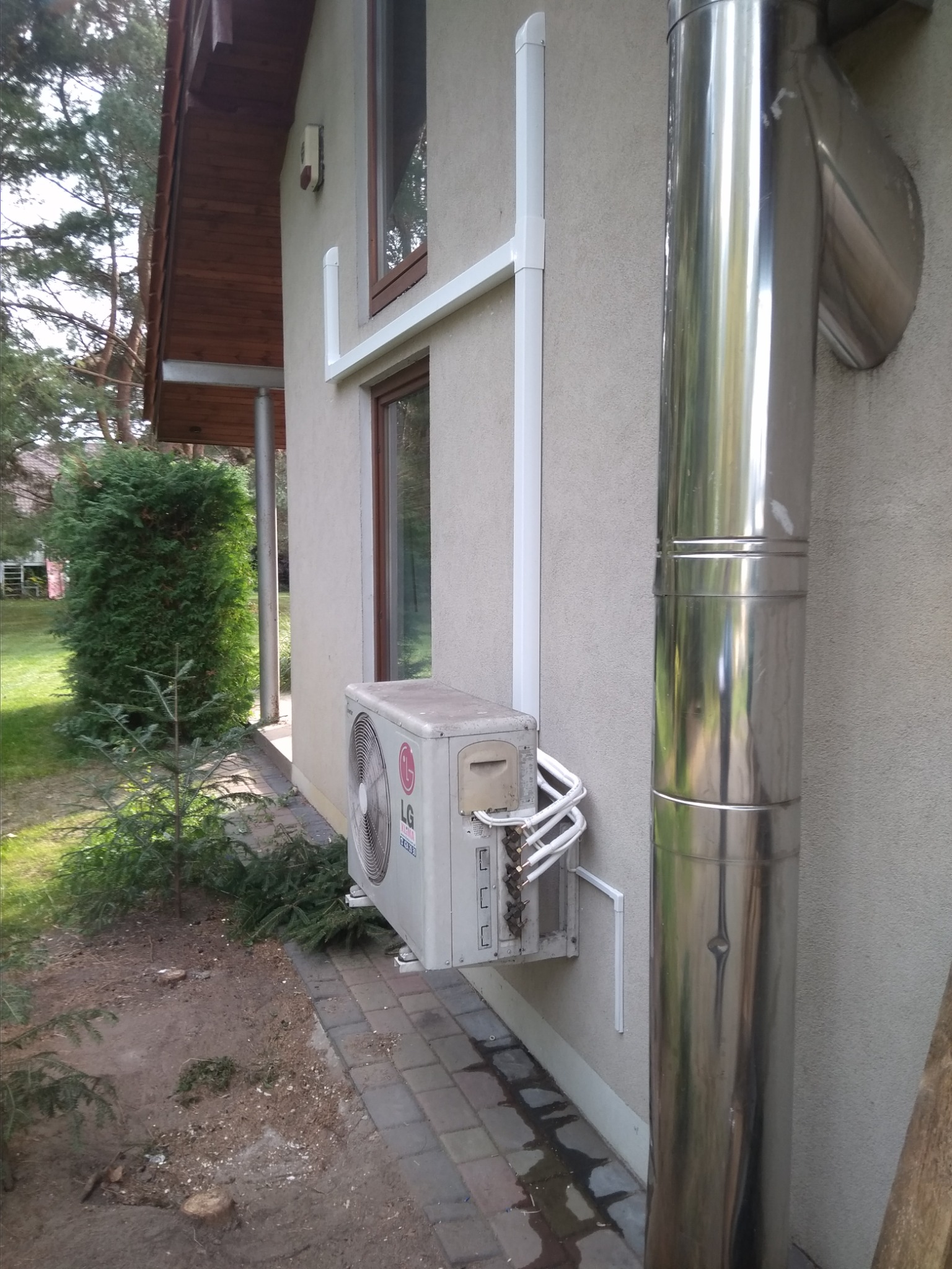 Zewnętrzna jednostka klimatyzacji LG zamontowana na elewacji domu, widoczne białe rurki instalacyjne i błyszczący, metalowy komin wentylacyjny obok.