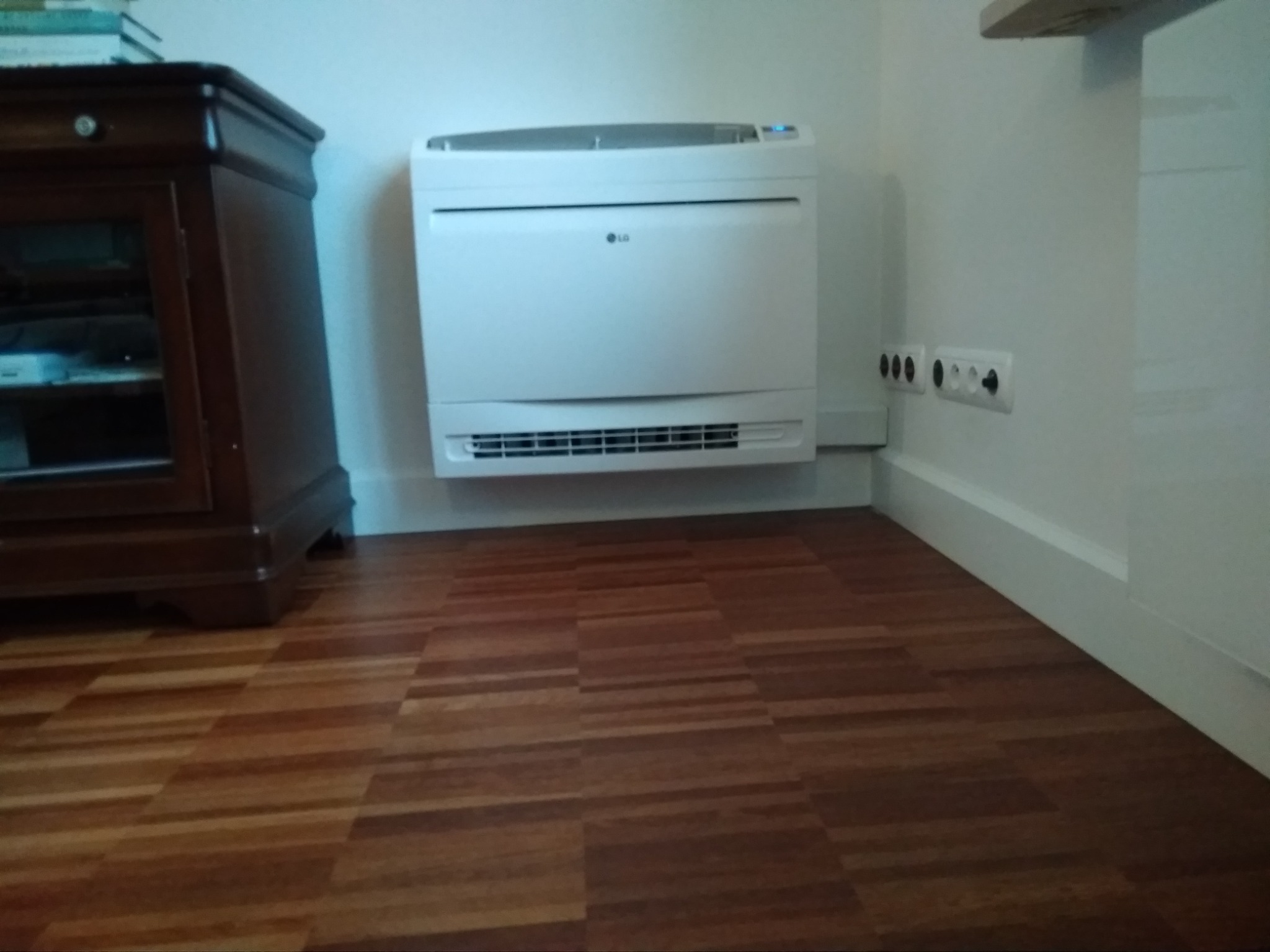 Wolnostojący klimatyzator LG w jasnym pomieszczeniu, widoczne gniazdka elektryczne i drewniana podłoga.