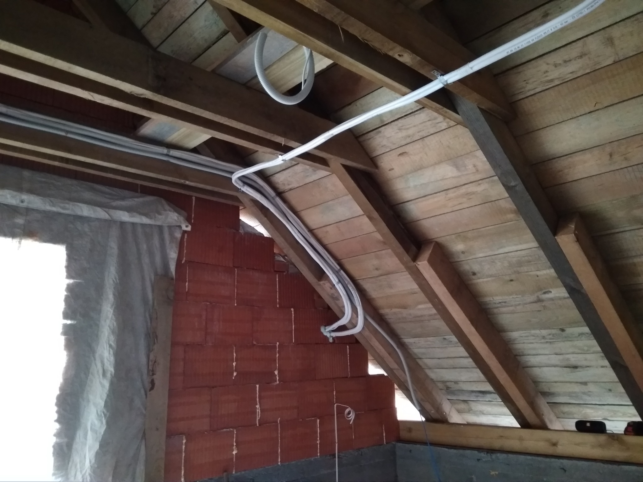 Instalacja przewodów wentylacyjnych w przestrzeni poddasza z drewnianą konstrukcją dachu i fragmentem ściany z czerwonej cegły.