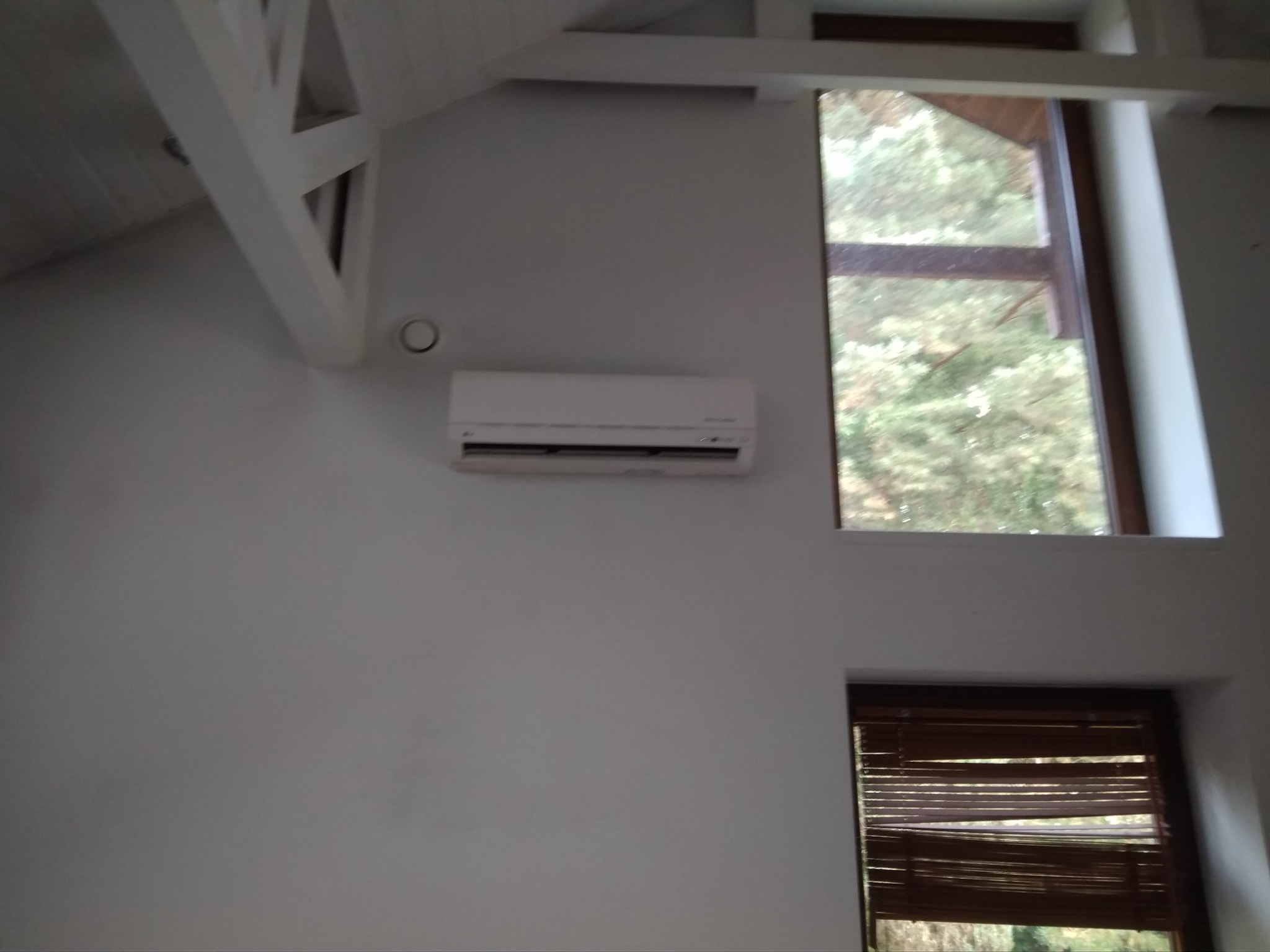 Biały klimatyzator ścienny zamontowany w pokoju z oknami i drewnianą konstrukcją sufitu, widok na zewnątrz przez okno.