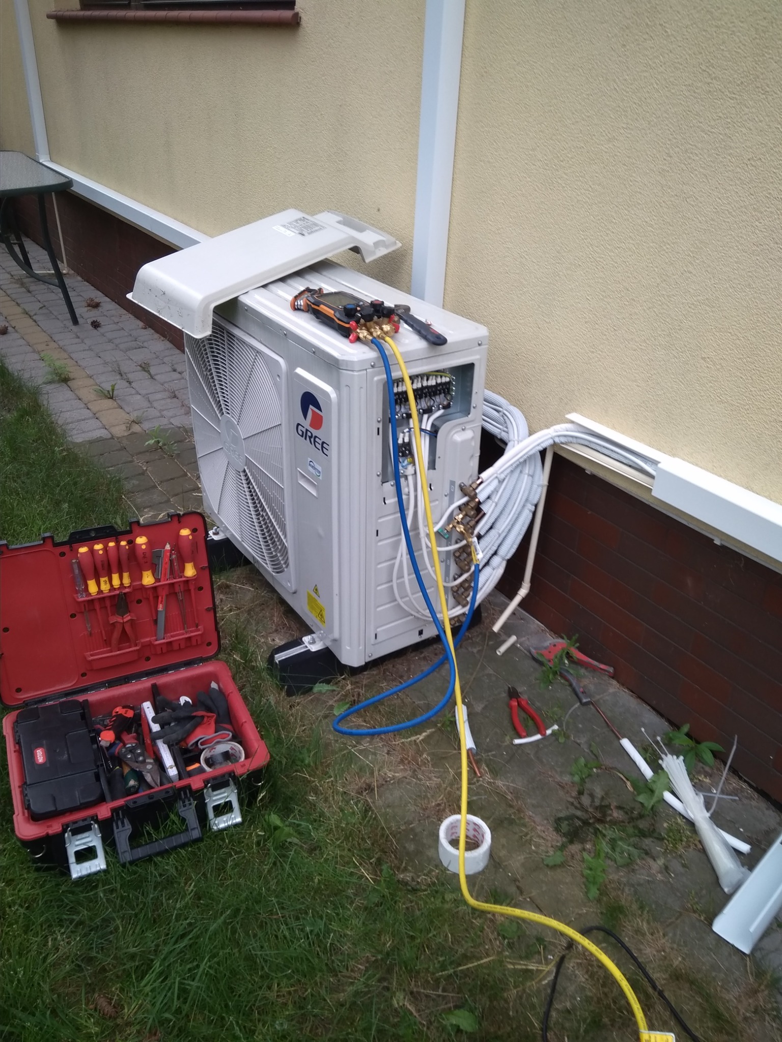 Jednostka zewnętrzna klimatyzatora marki Gree, z otwartą obudową, podłączona do przewodów, narzędzia pomiarowe na górze, obok otwarta skrzynka z narzędziami na trawie, widoczne elementy instalacji...